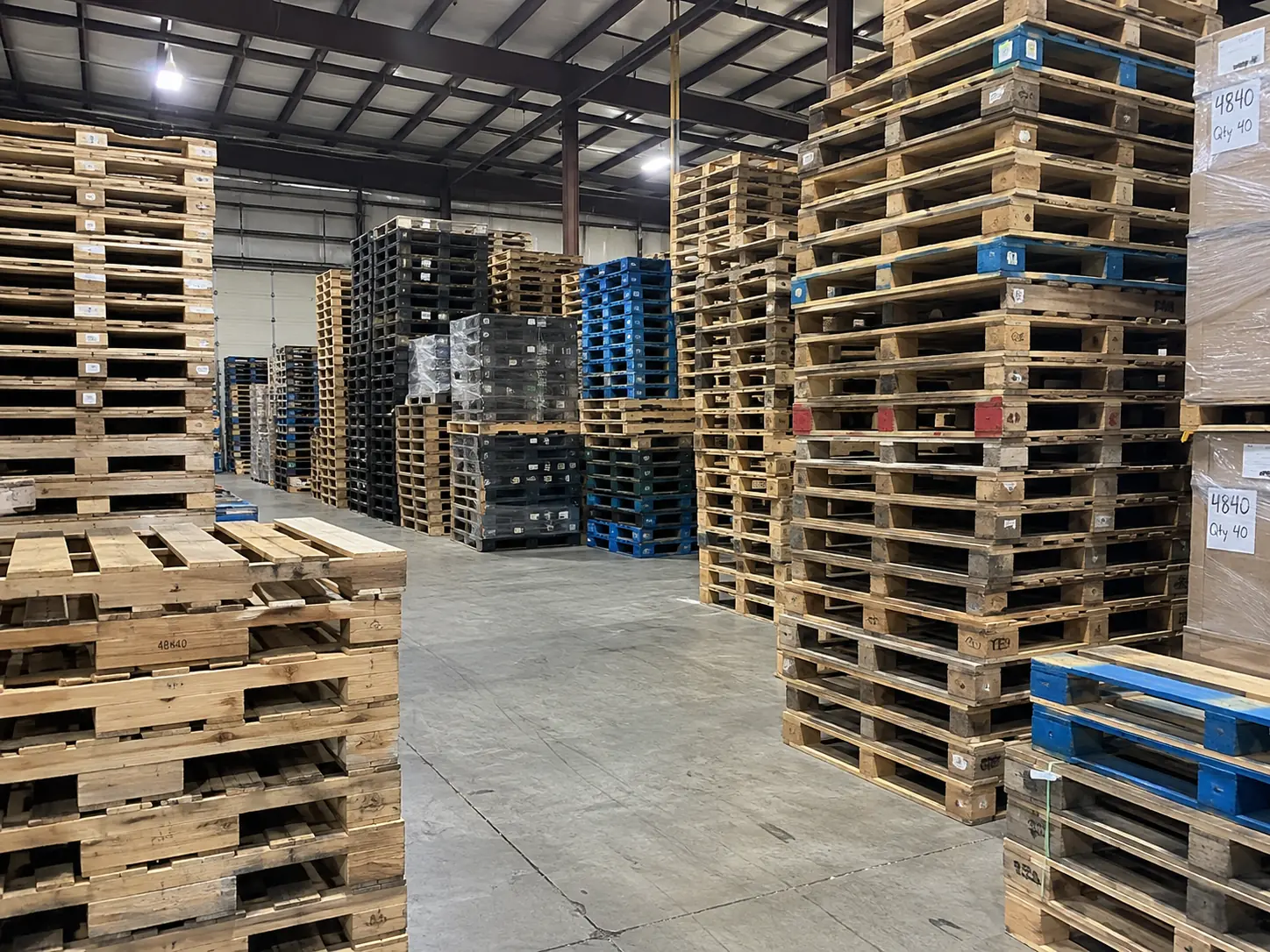 An aisle inside the Evendale warehouse, lined with banded stacks of recycled wood pallets carrying lot tags, alongside stacks of black and blue plastic pallets.